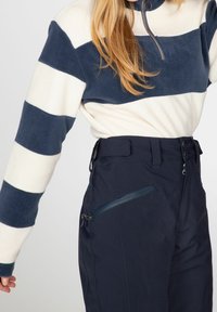 Pull en polaire à rayures horizontales bleu marine et crème, col zippé partiel ; associé à un pantalon bleu marine avec poche zippée et passants de ceinture.