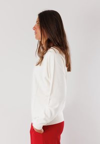 Maglione bianco a maniche lunghe con collo rotondo, caratterizzato da una texture morbida e una vestibilità leggermente rilassata, abbinato a pantaloni rossi.