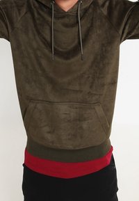 Felpa verde oliva realizzata in morbido tessuto, con tasca frontale, cordini regolabili e un elastico rosso contrastante in basso.