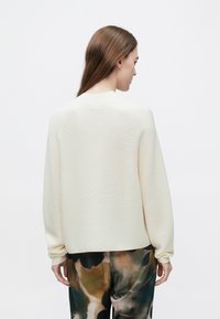 Kremowy sweter z teksturowanym splotem, prążkowanym dekoltem i długimi rękawami, zestawiony z luźnymi spodniami w wzory.