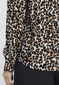 Blusa con stampa leopardata nei toni del nero, marrone e crema; presenta polsini a bottoni e un orlo curvo, realizzata in tessuto leggero e liscio.