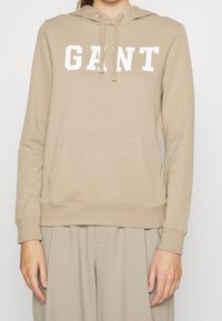 Béžová mikina vyrobená z měkké tkaniny s přední kapsou a bílým textem "GANT". Obsahuje kapuci na šňůrku a vroubkované cuffy.