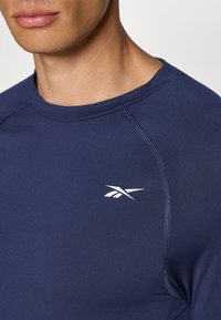 Cămașă sport de culoare albastru marin cu guler rotund, având un logo alb pe pieptul stâng și un material neted, elastic.