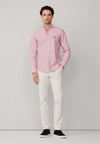 Camisa de botón a rayas rojas y blancas con cuello, combinada con pantalones beiges y zapatos oscuros. La camisa presenta un logo en el pecho.