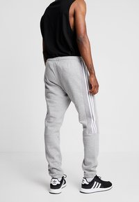 Man med grå joggingbyxor med vita ränder och svarta Adidas-sneakers, stående mot en enkel ljus bakgrund, vänd bort.