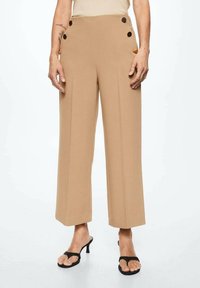 Beige wijde broek gemaakt van een gladde stof, met aan elke zijde twee decoratieve knopen, en gecombineerd met zwarte sandalen.