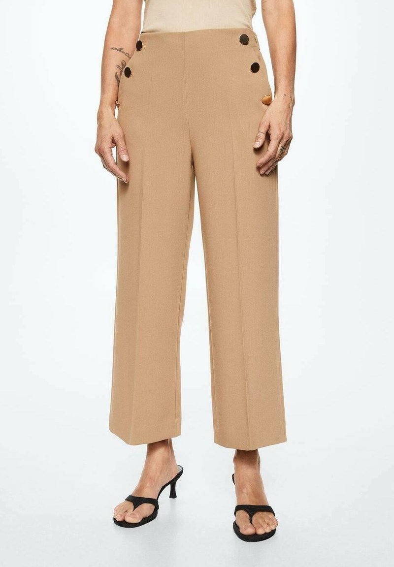 Beige wijde broek gemaakt van een gladde stof, met aan elke zijde twee decoratieve knopen, en gecombineerd met zwarte sandalen.