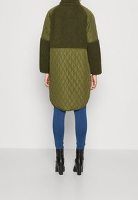 Person med en kjol längd olivgrön vadderad och fleecejacka, blå skinny jeans och svarta klackade ankle boots, visad bakifrån.