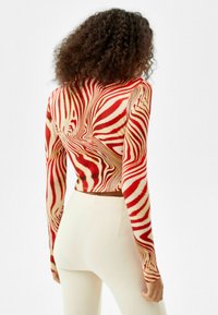 Haut court à manches longues avec un motif tourbillonnant rouge et crème. Confectionné dans un matériau ajusté et extensible. Porté avec un pantalon crème moulant.