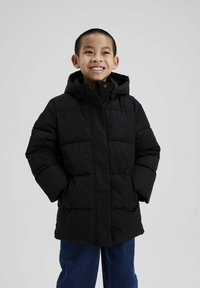 Schwarze Daunenjacke mit Kapuze, mit gestepptem Design, Frontreißverschluss und Seitentaschen. Getragen über Jeans.