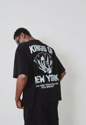 Homme portant des lunettes de soleil noires et un T-shirt noir avec l'inscription "Kings of New York" et des motifs de cartes à jouer dans le dos, sur un fond uni.