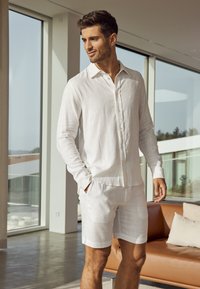 Camicia bianca in lino a maniche lunghe con colletto button-down, abbinata a pantaloni corti coordinati. Texture liscia, design leggero e vestibilità rilassata.
