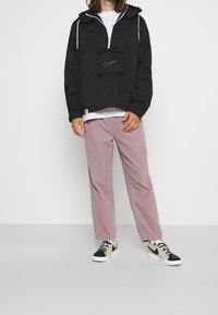 Person wearing a black Nike hoodie, white t-shirt, mauve pants, and white and black Nike sneakers standing against a plain background.