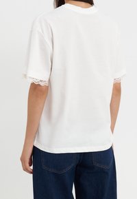 Osoba ubrana w białą koszulę z krótkim rękawem i koronkowymi wykończeniami na rękawach, w niebieskie dżinsy, pokazana z tyłu na jednolitym tle.