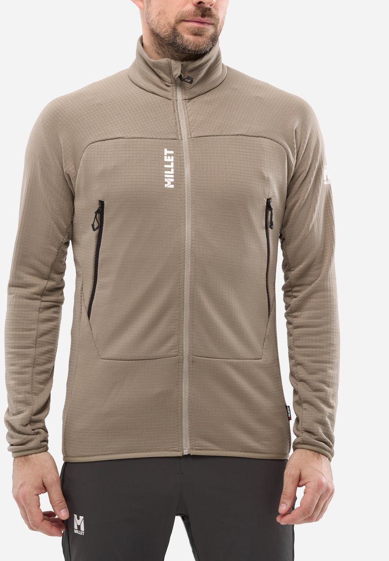 Un homme portant une veste en polaire beige à fermeture éclair avec des poches zippées noires et un pantalon noir, arborant des logos "Millet" sur la veste et le pantalon.