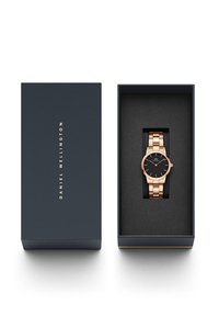 Gold-tone metal watch with a black face, linked bracelet, displayed in a navy blue box lined with black fabric. Brand name on the box.