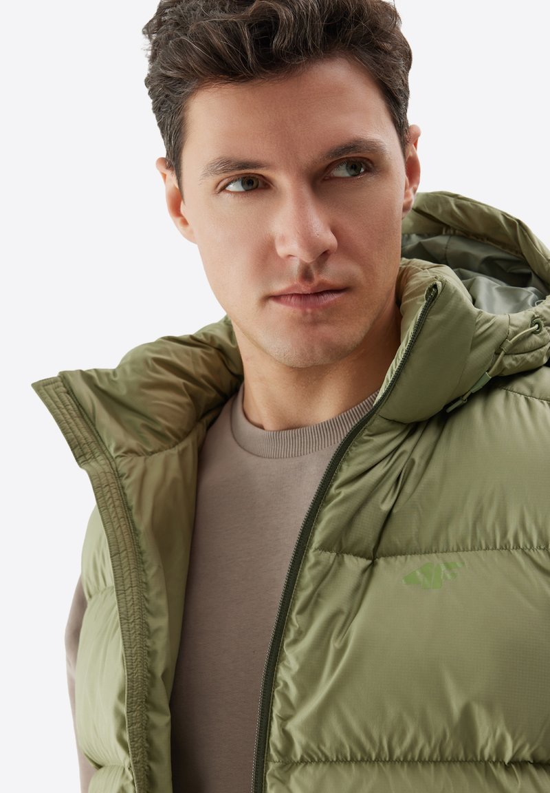 Oliwkowa puchowa kurtka z wysokim kołnierzem i zapięciem na zamek, charakteryzująca się pikowaną teksturą i subtelnym logo na klatce piersiowej. Noszona na beżowej koszuli.