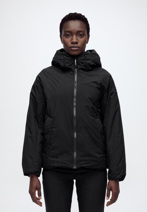 Une femme porte une veste noire zippée à grande capuche et un pantalon noir sur un fond blanc uni.