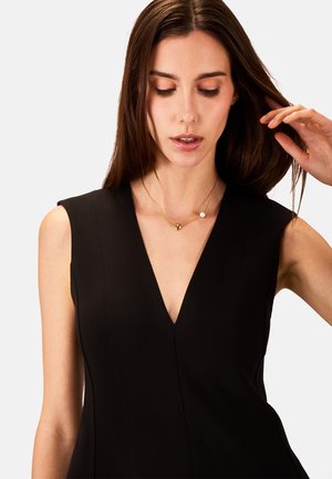 Abito nero con scollatura a V profonda e silhouette aderente. Presenta delicate cuciture verticali e design senza maniche. Collana in oro e perle in accento.