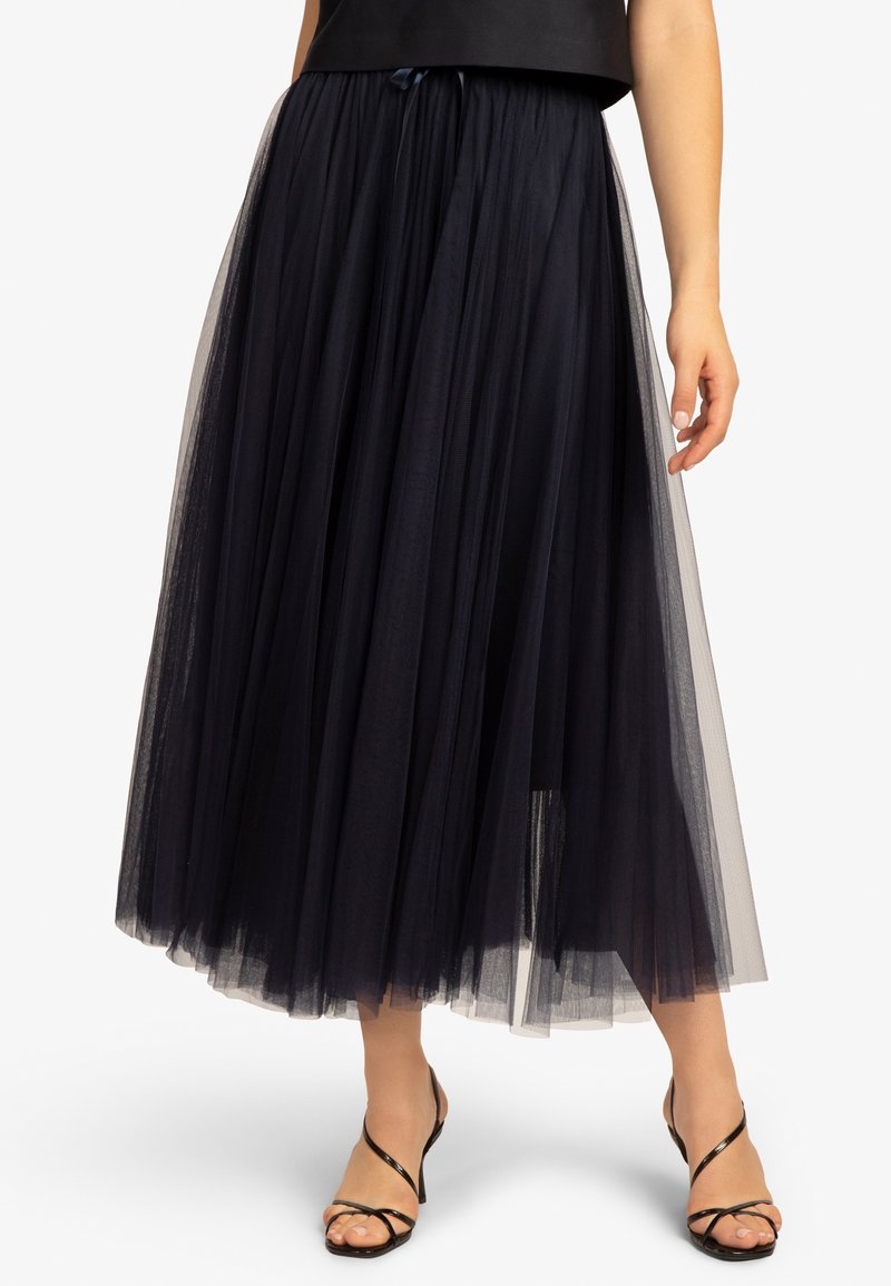 Jupe midi en tulle noir superposée, portée avec des sandales à talons hauts noires à lanières, laissant voir les jambes à partir du milieu de la cuisse vers le bas.