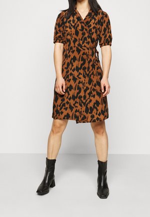 Kobieta w brązowo-czarnej sukience kopertowej w zwierzęcy wzór, w połączeniu z czarnymi botkami na obcasie, stojąca na jednolitym jasnym tle.