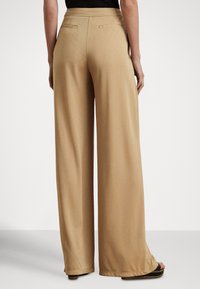 Beżowe spodnie o szerokich nogawkach o gładkiej fakturze, z tylnymi kieszeniami i wysokim stanem, zestawione z czarnymi sandałami na platformie.