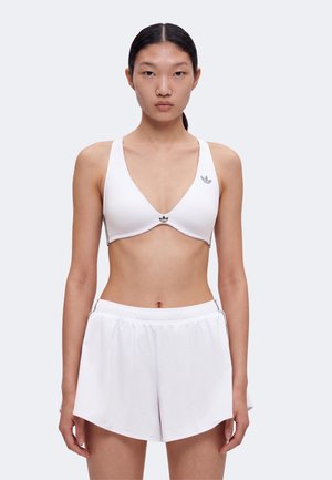 Kvinde stående med front mod kamera iført hvid sports-bh og hvide sportsshorts mod ensfarvet baggrund.