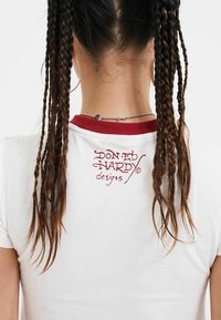 Hvid t-shirt med en rød ribbet krave, der har et trykt logo "DON ED HARDY designs" på den øverste ryg. Korte ærmer, glat tekstur.