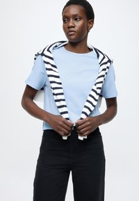 Ανοιχτό μπλε βαμβακερό μπλουζάκι με στρογγυλή λαιμόκοψη, σε συνδυασμό με μαύρο τζιν. Μια μπλε ναυτική και λευκή ριγέ ζακέτα είναι περασμένη στους ώμους.