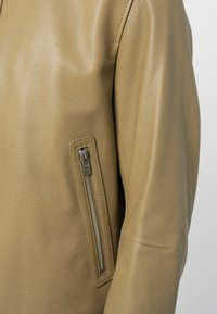 Κοντινό πλάνο μανικιού κάμελ δερμάτινου μπουφάν με κάθετη ασημί τσέπη με φερμουάρ και εμφανείς λεπτομέρειες ραφών.