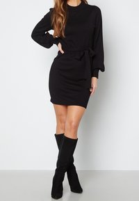 Robe noire à manches longues avec une silhouette ajustée, une texture côtelée, et une ceinture à la taille. Associée à des bottes en suède noir montant jusqu'aux genoux.