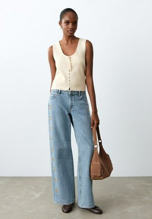 Mujer con top beige sin mangas y pantalones vaqueros de pierna ancha azul con bordado floral, sosteniendo un bolso marrón de hombro, de pie sobre un suelo gris.