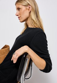 Femme blonde dans un pull noir, s'appuyant sur une chaise tressée avec accoudoirs en métal noir, regardant à gauche avec une expression relaxée.
