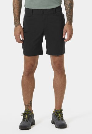 Mężczyzna w czarnych szortach i szarych butach trekkingowych z żółtymi detalami, stojący na jednolitym jasnym tle.