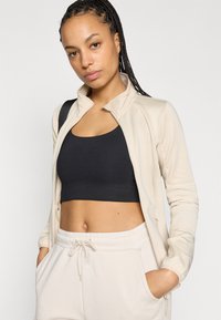 Veste beige à fermeture éclair avec un col montant, portée sur un haut court noir. Présente une texture lisse et une coupe ajustée avec des poches latérales.