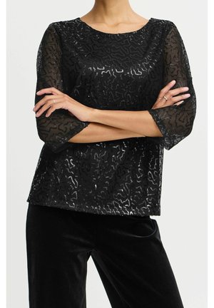 Zwarte doorschijnende top met driekwartmouwen, voorzien van een textuurpatroon en verspreide pailletten. Genieuwd met zwarte fluwelen broek.