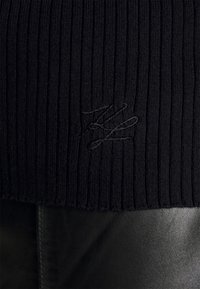 Svart ribbad stickad tröja med en subtil broderad logotyp. Texturen är mjuk, med en kroppsnära passform, ihop med en slät del av svart läder.