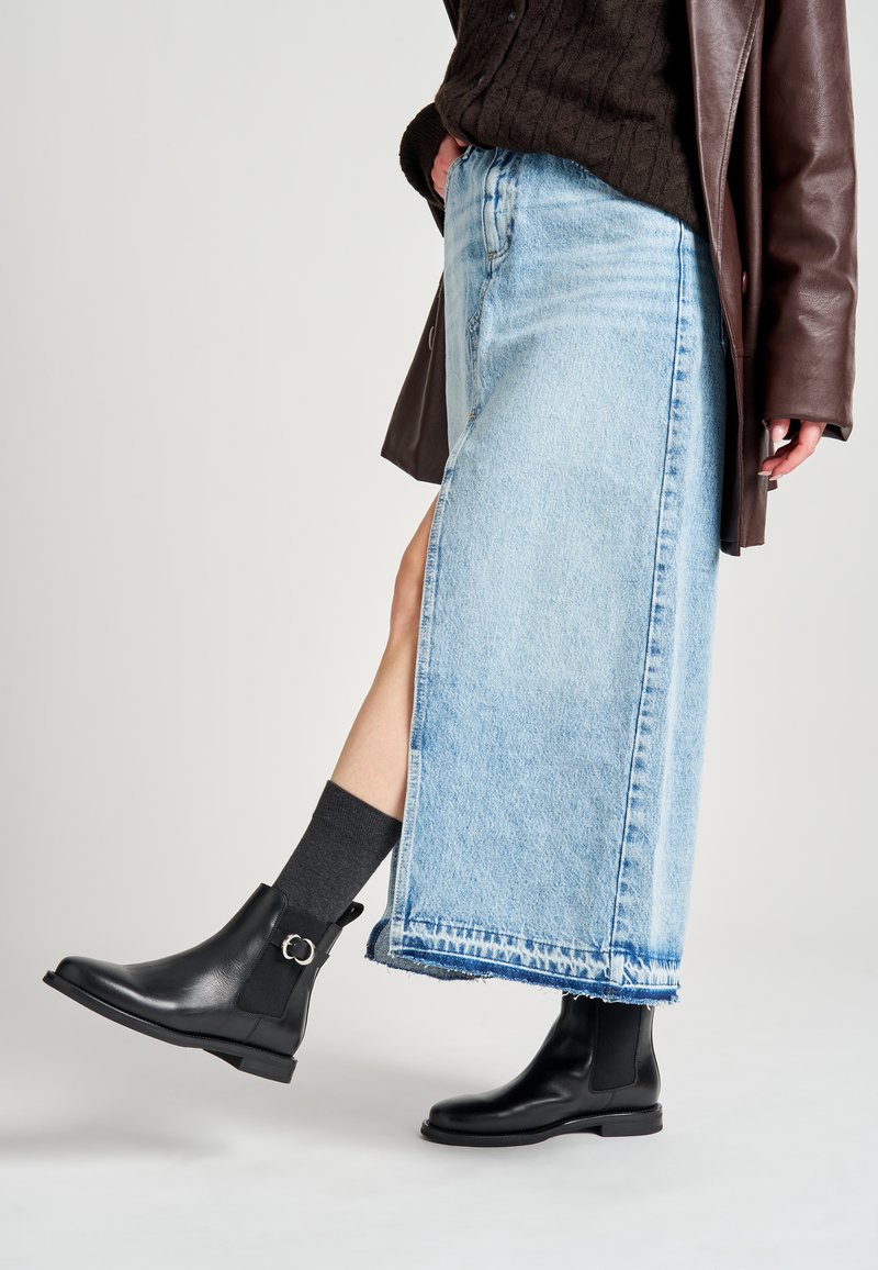 Botas de cuero negras hasta el tobillo con punta redonda y laterales elásticos, combinadas con una falda de mezclilla azul clara que presenta un dobladillo deshilachado y una abertura lateral.