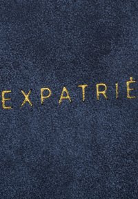 Tengerészkék zamak anyag, melyen az "EXPATRIÉ" szó fényes aranyszínűen van beleégetve, sima textúrával és minimális dizájnnal.