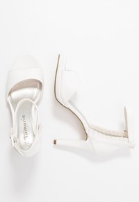 Bílé sandály na vysokém podpatku s otevřenou špičkou, páskem přes přední část a texturovaným zadním dílem. Mají úzký podpatek a lesklý povrch.