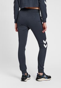 Mörkblå sweatpants med en avsmalnande design, som har vita chevron-accenter på sidorna och ribbade muddar, tillsammans med svarta träningsskor.