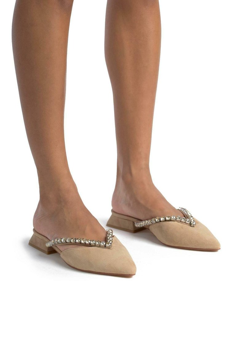 Mules en suède beige avec un embout pointu et un petit talon, dotées d'une sangle en forme de V accentuée de cristaux sur le coup de pied.