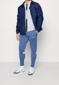 Navy windbreaker over a gray T-shirt with black stripes, paired with blue athletic leggings and white sneakers with neon green accents.