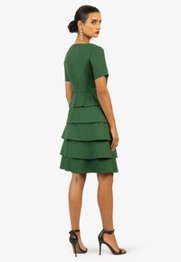 Grünes gestuftes Kleid mit kurzen Ärmeln, strukturiertem Stoff, tailliertem Oberteil und ausgestellt geschichtetem Rock. Die Modell trägt schwarze High Heels.