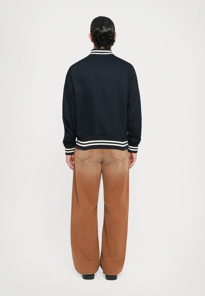 Veste bombardier bleue marine avec des poignets et une taille en côtes rayées blanches, associée à un pantalon en jean large marron clair, vue de dos.