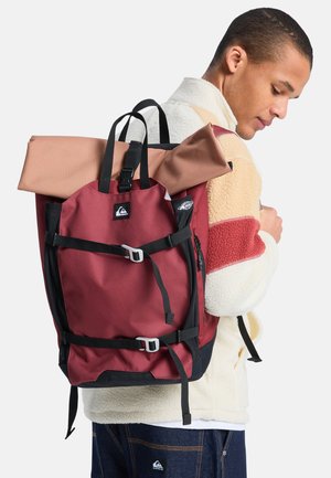 Młody mężczyzna w kremowo-czerwonej polarowej kurtce niesie duży bordowy plecak z czarnymi paskami i zwiniętym materacem na górze.