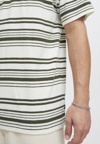 Høyre arm til person med hvit og mørkegrønn stripete skjorte og sølvkjede håndleddsarmbånd, beige bukser synlige nederst.