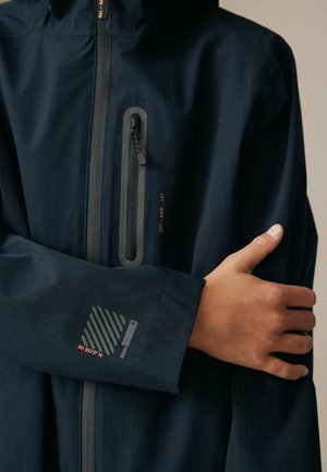 Person, die eine dunkelmarineblaue Jacke mit vertikalem Brust-Reißverschlusstaschen und gedruckten Koordinaten am Ärmel trägt, die Arme vor der Brust verschränkt.