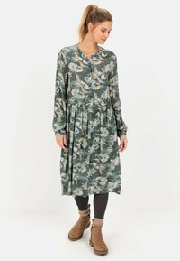 Kvinde med blond hestehale iført en grøn og beige blomsterlangærmet midikjole, sorte leggings og brune ankelstøvler med beige sokker.