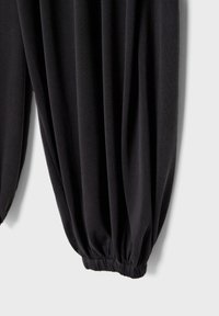 Pantalon en tissu noir avec un bas élastique, froncé et légèrement drapé sur un fond clair uni.